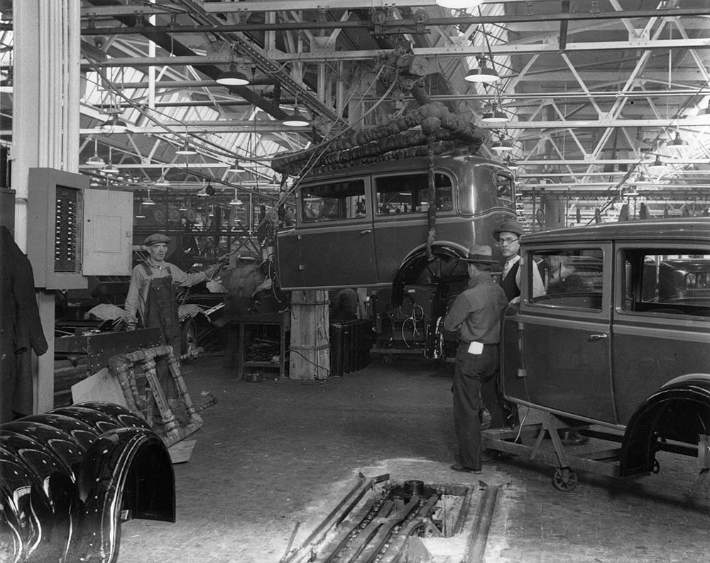 Interior view of unidentified car assembly plant, showing assembly line works, chassis, and workers