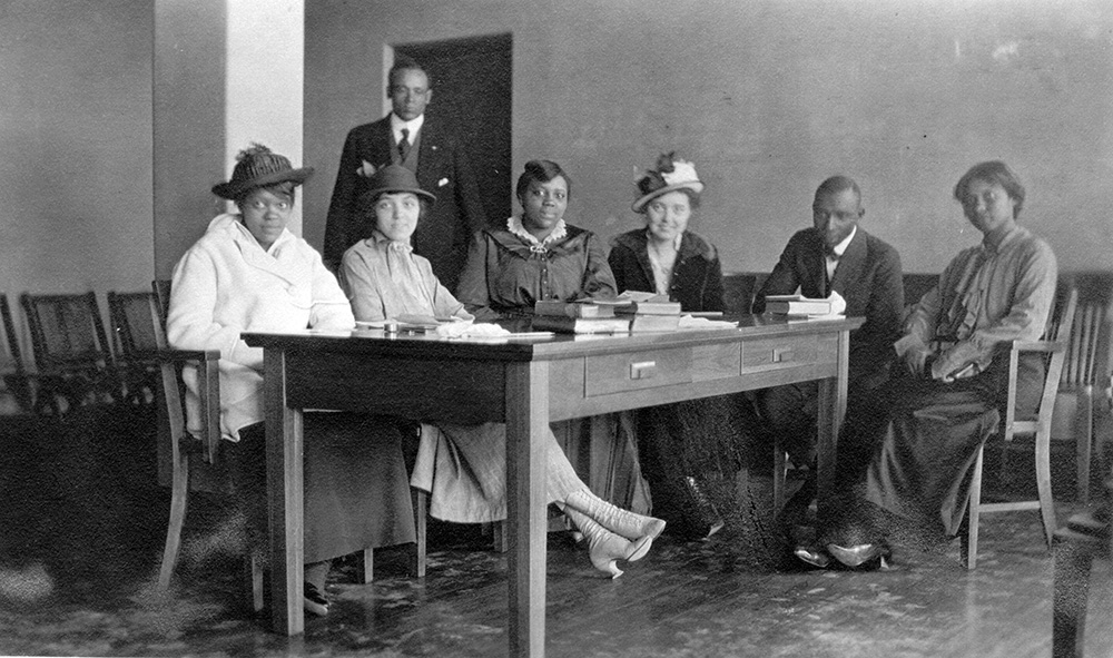 Six people sitting at a table full of books at a library, while a man stands in the background.