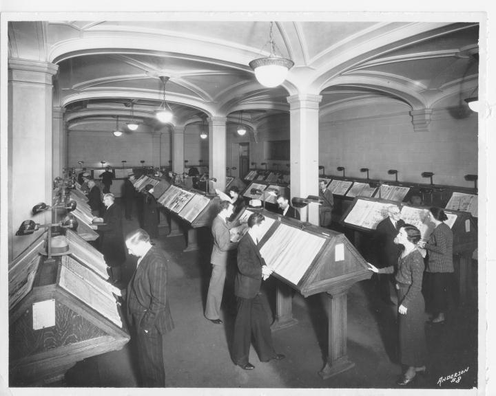 Library patrons reading newspapers on stands in a reading room