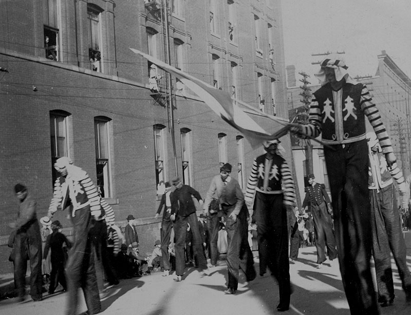 View of men on stilts in various dress walking along unidentified parade route during the Priests of Pallas parade