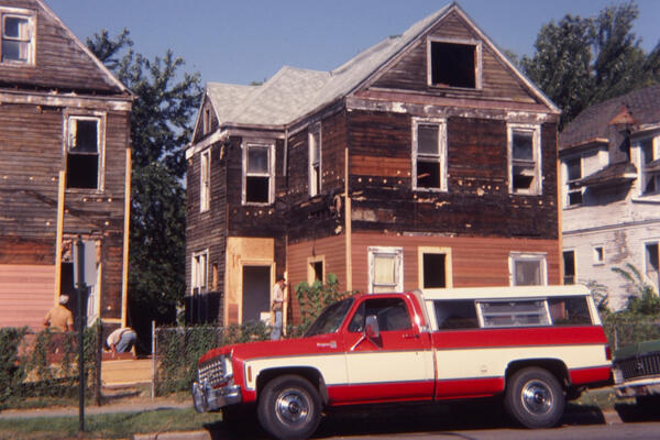 Color photo of a red and white truck outside a dilapidated house