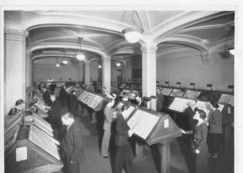 Library patrons reading newspapers on stands in a reading room