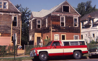 Color photo of a red and white truck outside a dilapidated house